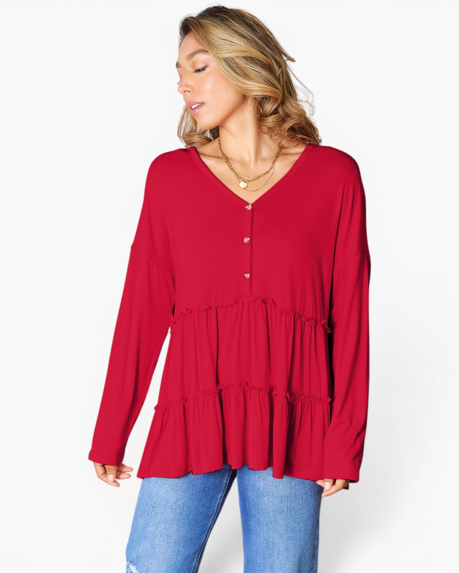 A woman wearing a red long-sleeve tiered blouse with a buttoned V-neck, paired with blue jeans, photographed against a plain white studio background.