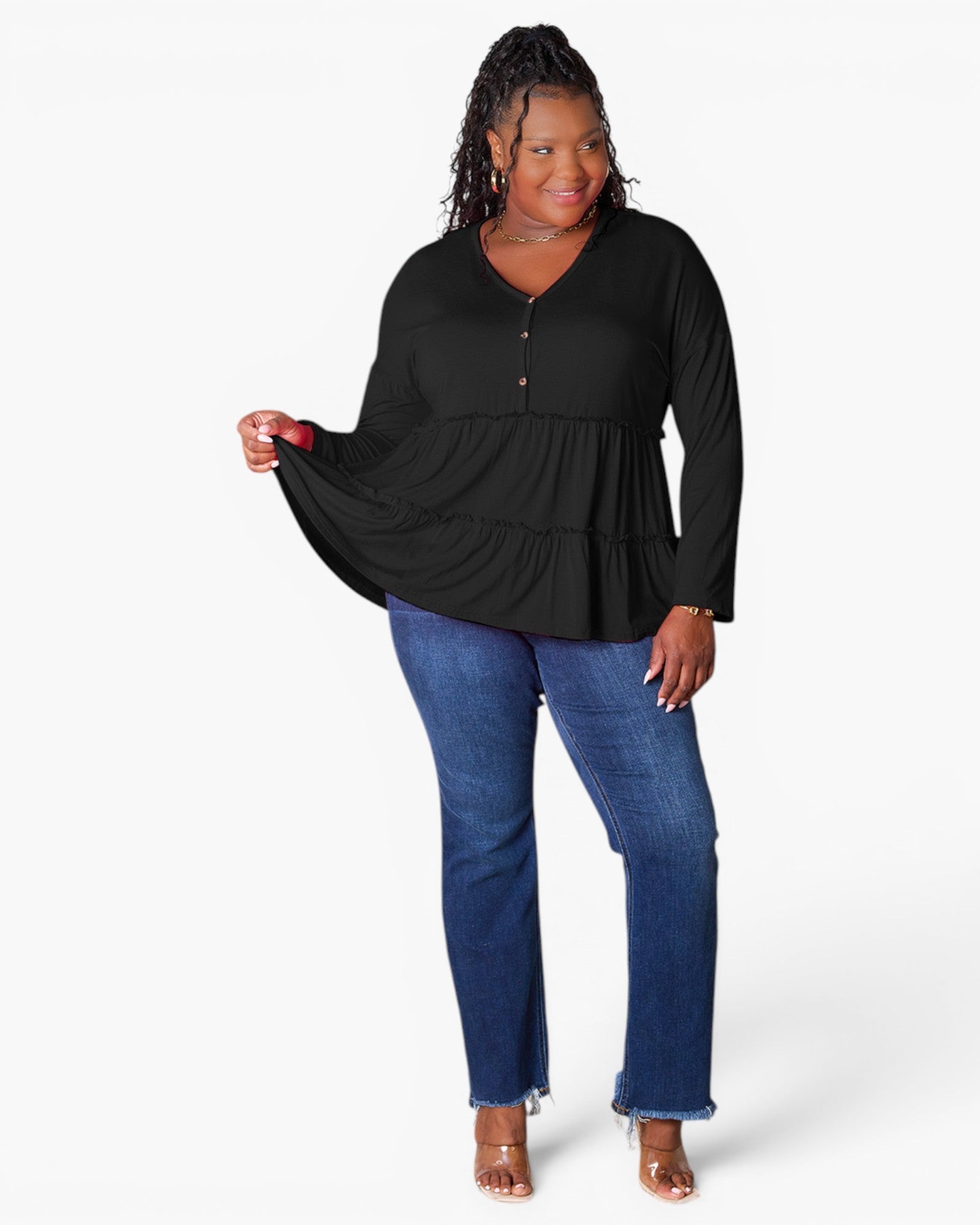 A woman modeling a black long-sleeve peplum top with a buttoned V-neckline, paired with blue jeans and clear high-heeled sandals, against a plain white studio background.