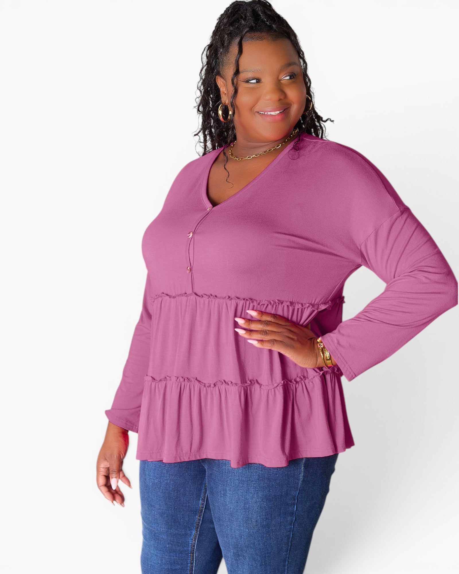 A woman modeling a long-sleeve V-neck top in dusty pink with a tiered peplum hem, paired with blue jeans, against a plain white studio background.