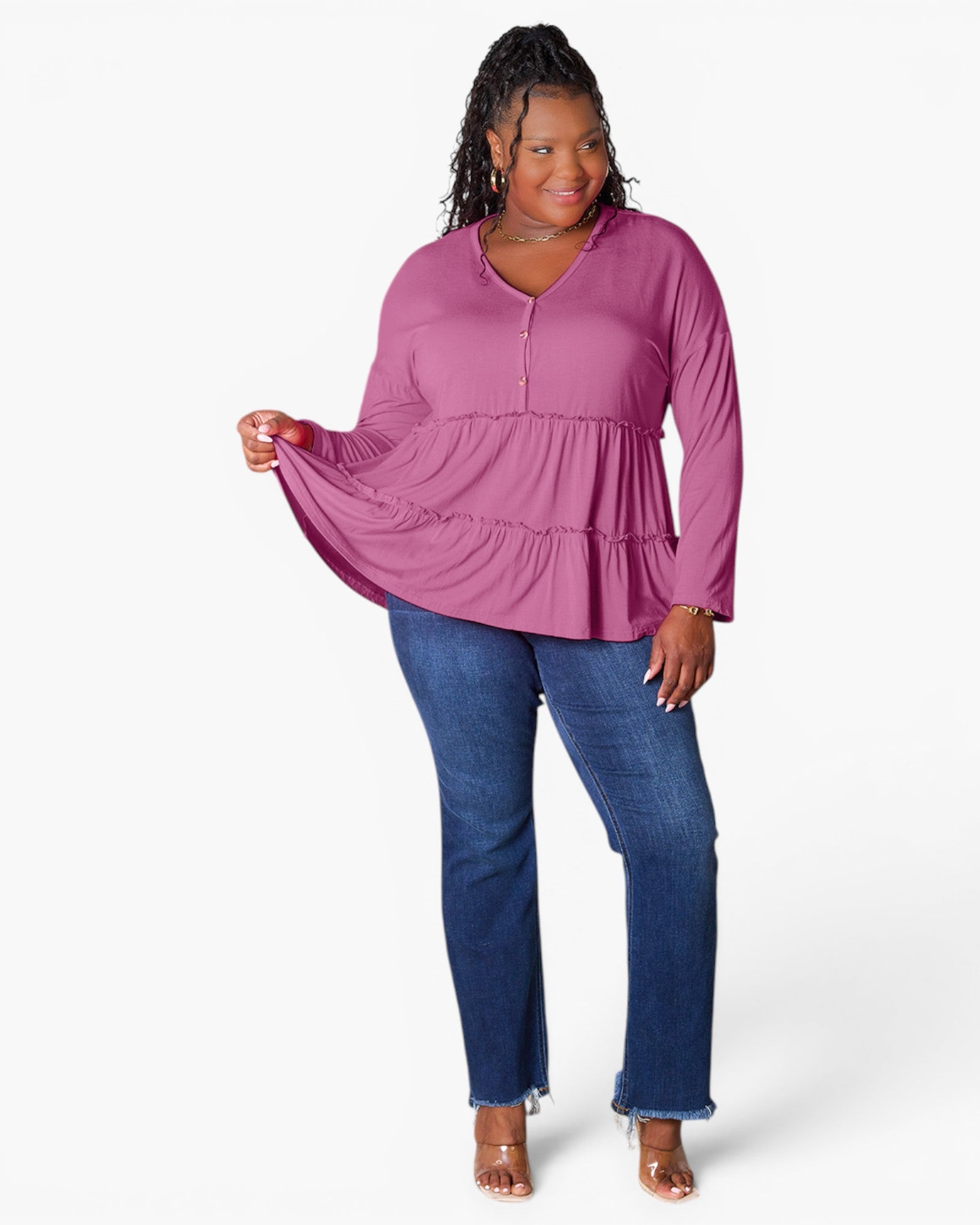 A woman modeling a long-sleeve tiered blouse in a dusty magenta-purple color, paired with blue jeans and clear high-heeled sandals, against a plain white studio background.