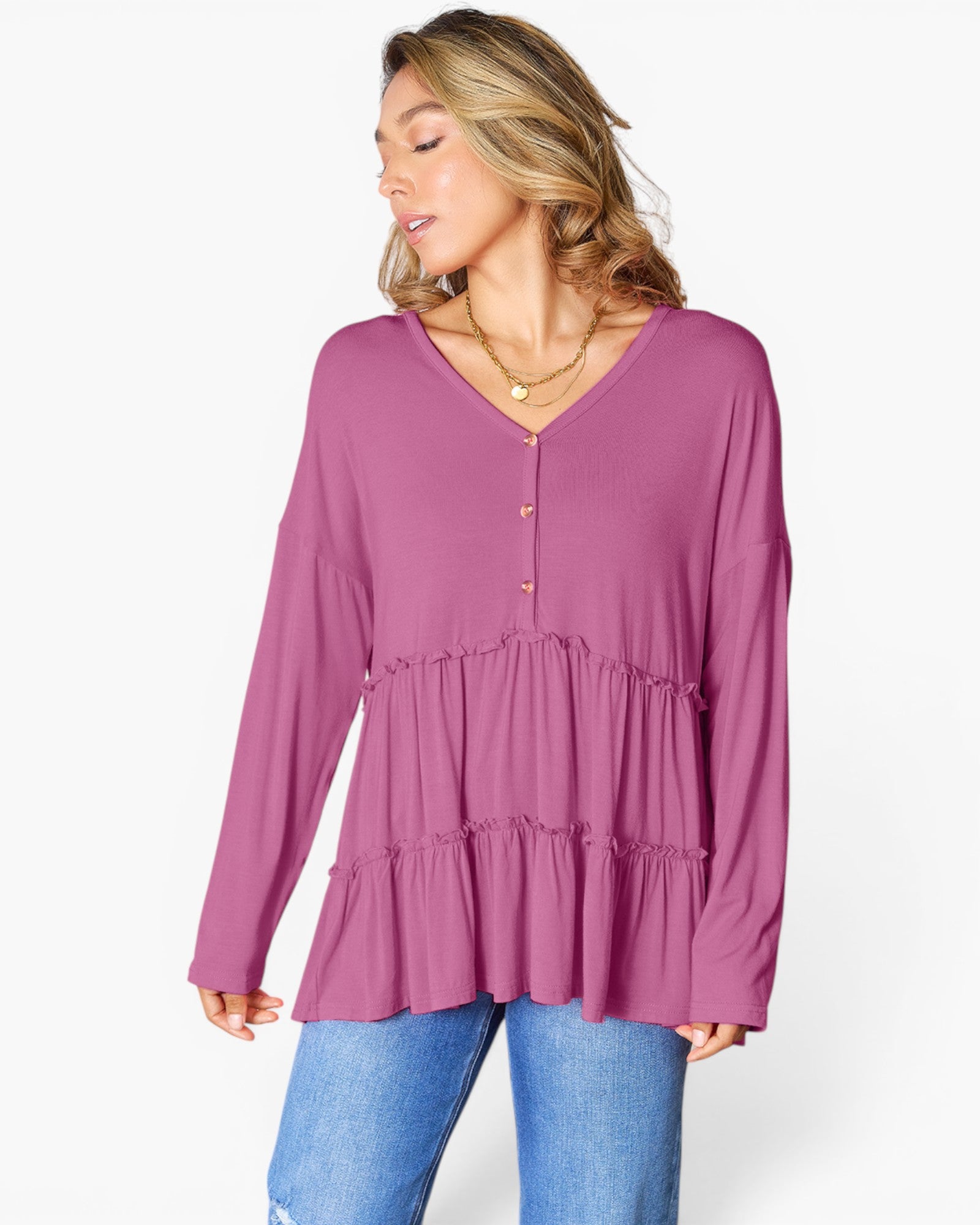 A woman wearing a dusty purple long-sleeve V-neck top with a tiered ruffle hem and button details, paired with blue jeans, against a plain white background.