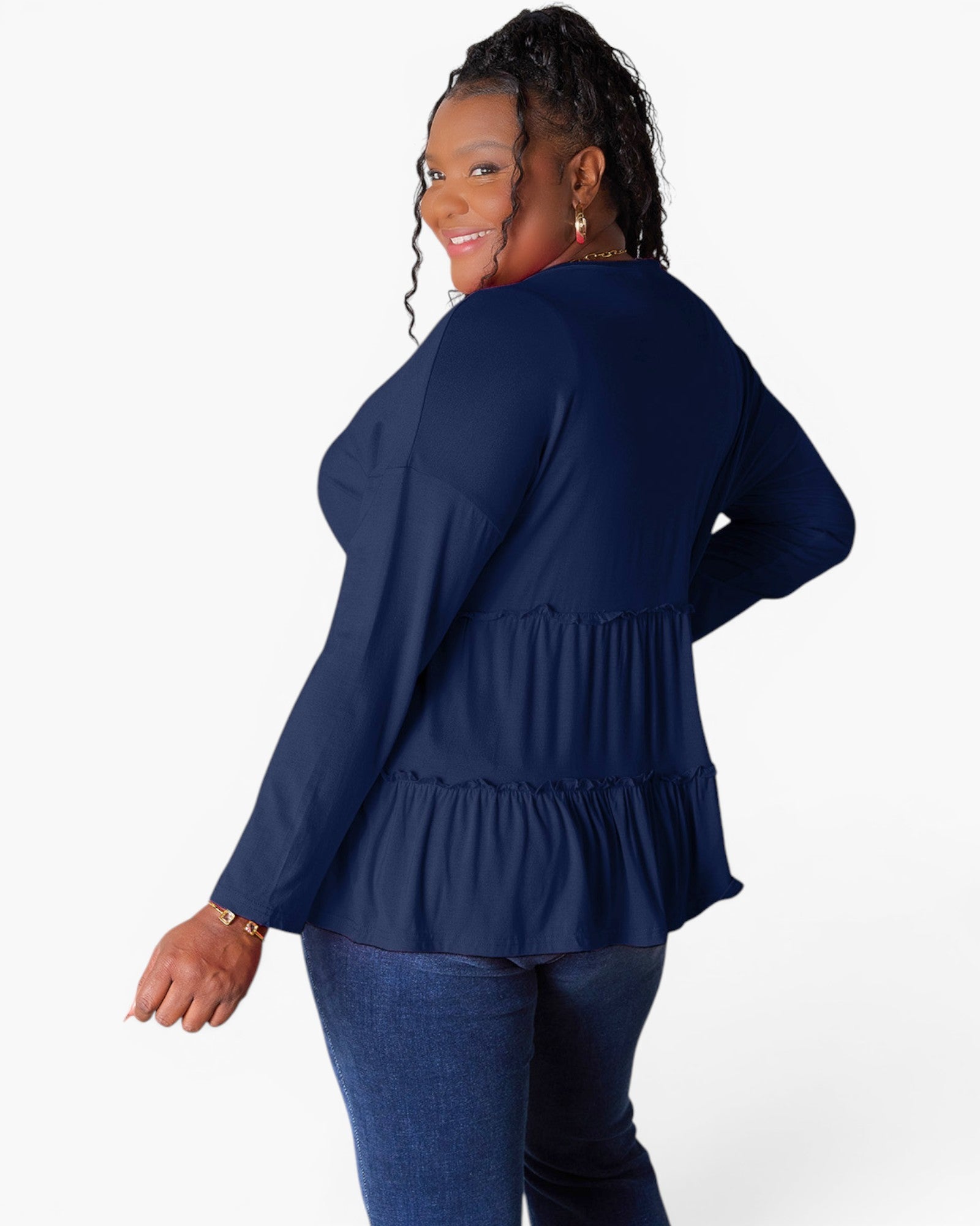 A woman modeling a navy blue long-sleeve top with a tiered ruffle hem, shown from a back-three-quarter angle against a plain white studio background.