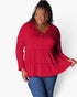 A woman modeling a red long-sleeve tiered blouse with a buttoned V-neckline, paired with blue jeans, against a plain white studio background.