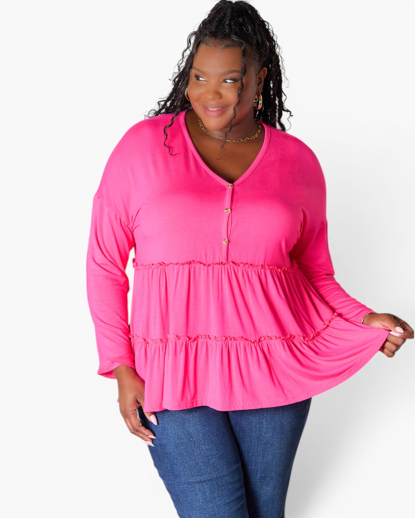 A woman modeling a bright pink long-sleeve tiered blouse with a buttoned V-neckline, paired with blue jeans, against a plain white studio background.