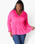 A woman modeling a bright pink long-sleeve tiered blouse with a buttoned V-neckline, paired with blue jeans, against a plain white studio background.