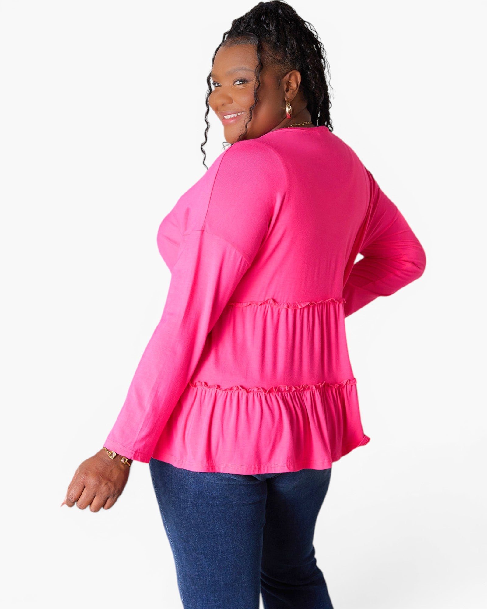 A woman modeling a bright pink long-sleeve top with a tiered ruffle hem, shown from a back-three-quarter angle against a plain white studio background.
