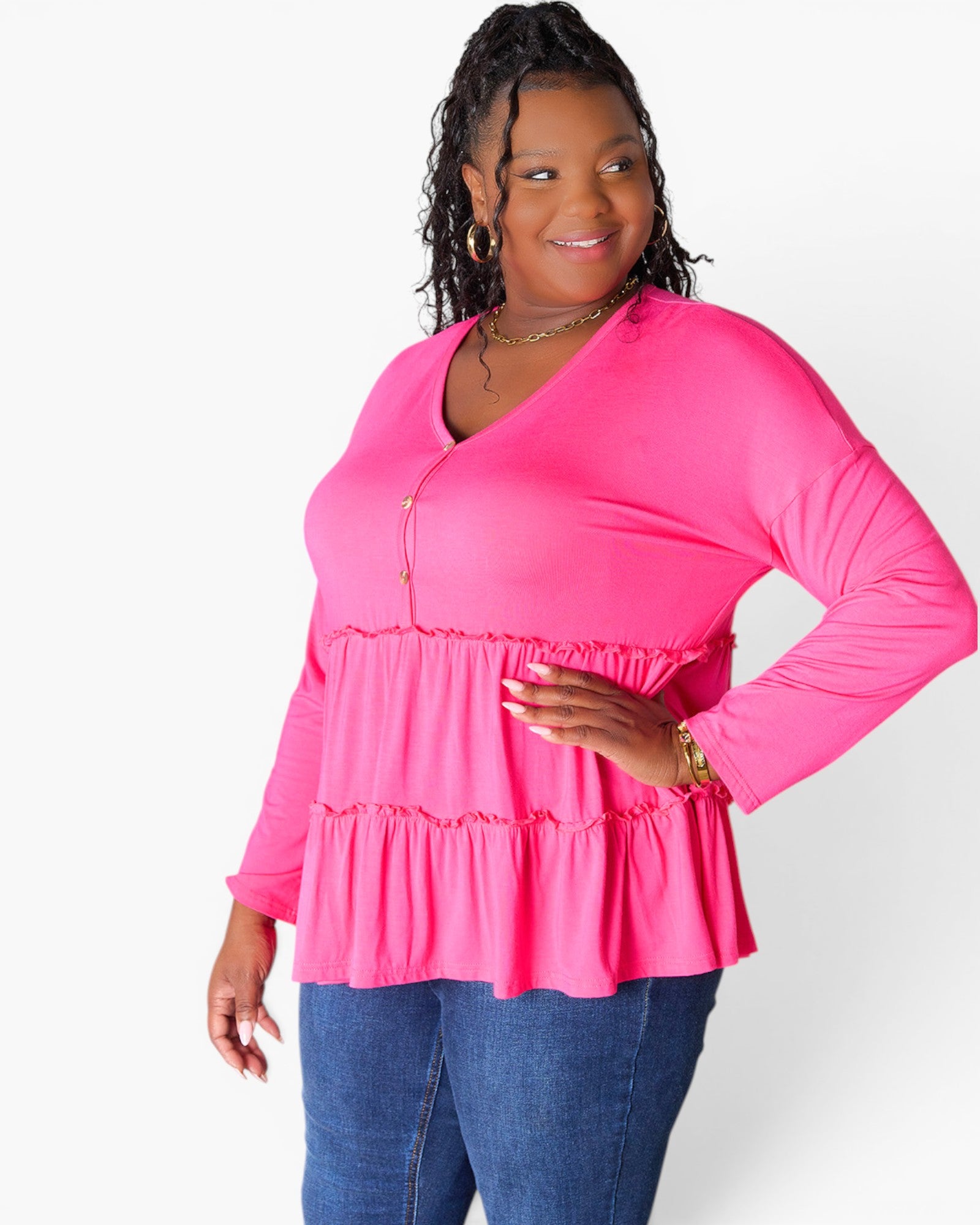 A woman modeling a bright pink long-sleeve top with a V-neck and tiered ruffle hem, paired with blue jeans, against a plain white studio background.