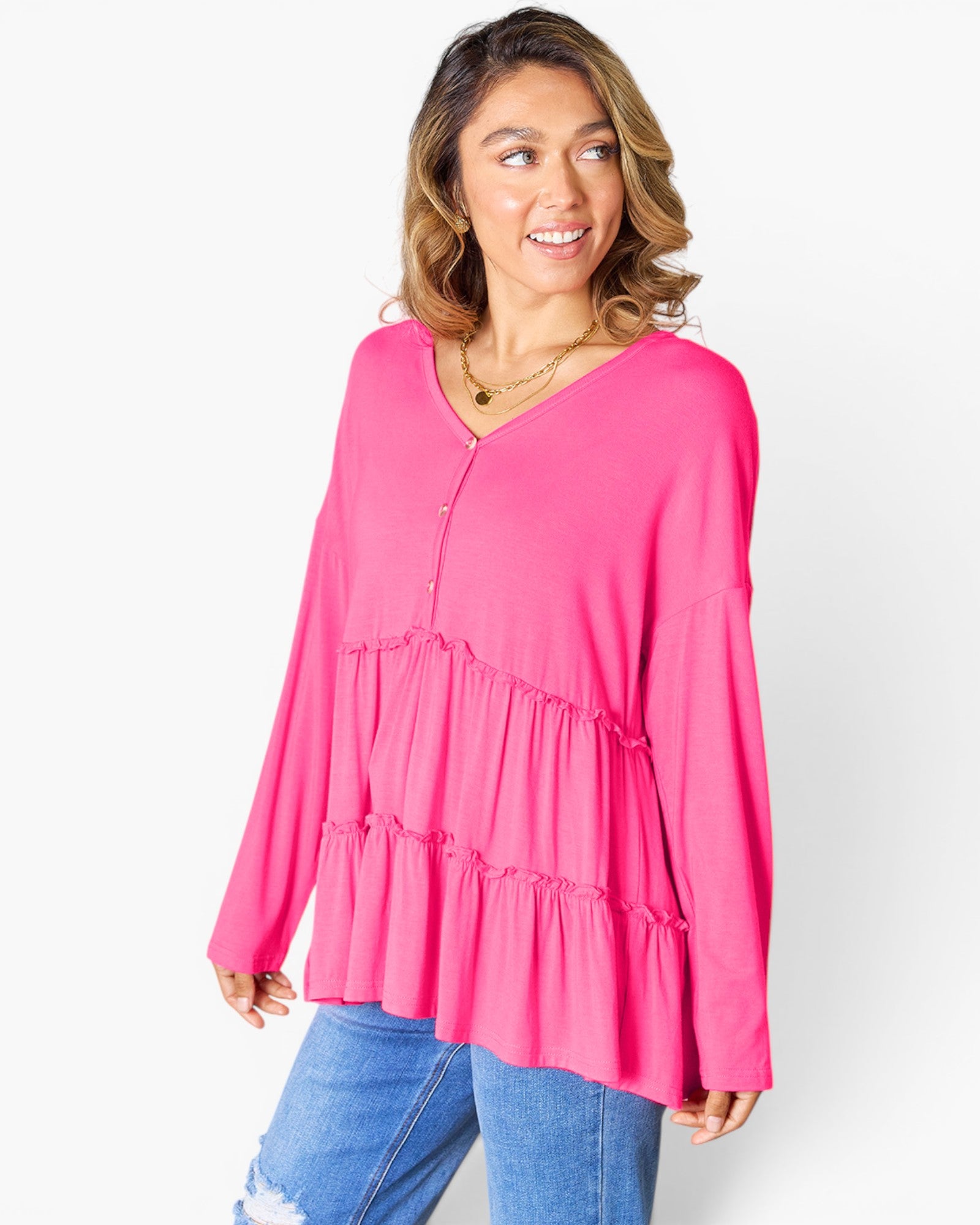 A woman wearing a bright pink tiered long-sleeve top with a buttoned V-neckline, paired with blue jeans, photographed against a clean white studio background.
