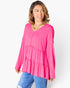 A woman wearing a bright pink tiered long-sleeve top with a buttoned V-neckline, paired with blue jeans, photographed against a clean white studio background.