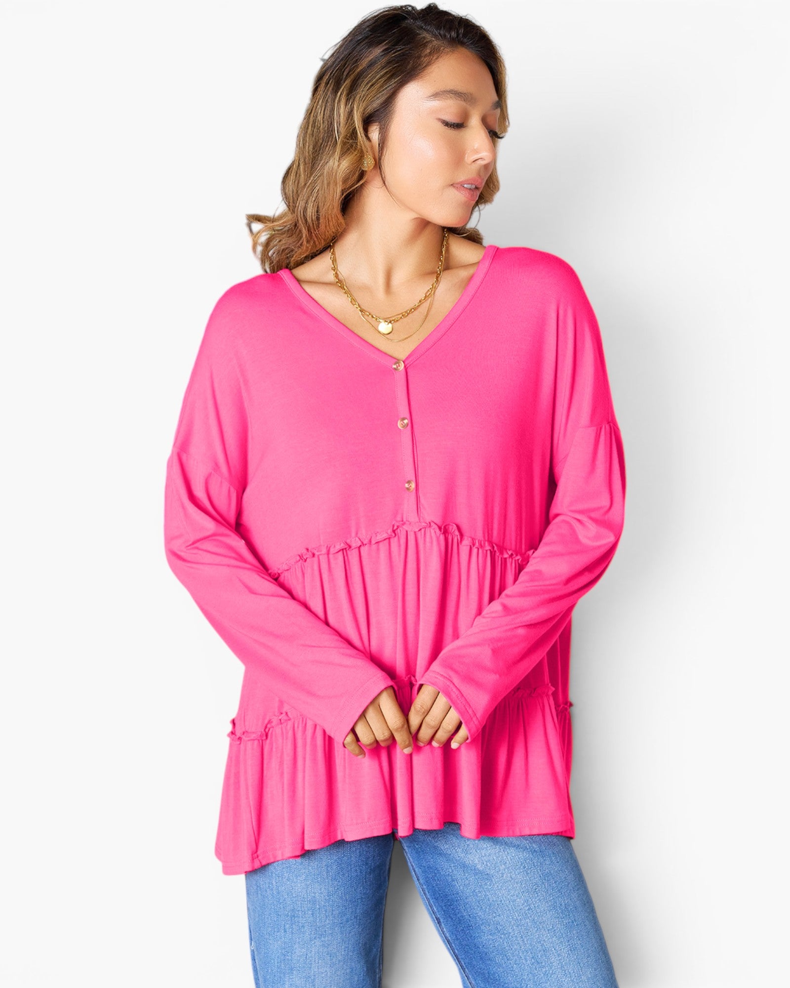 A woman wearing a bright pink long-sleeve V-neck top with a button placket and gathered ruffle detail at the waist, paired with blue jeans, against a light gray studio background.