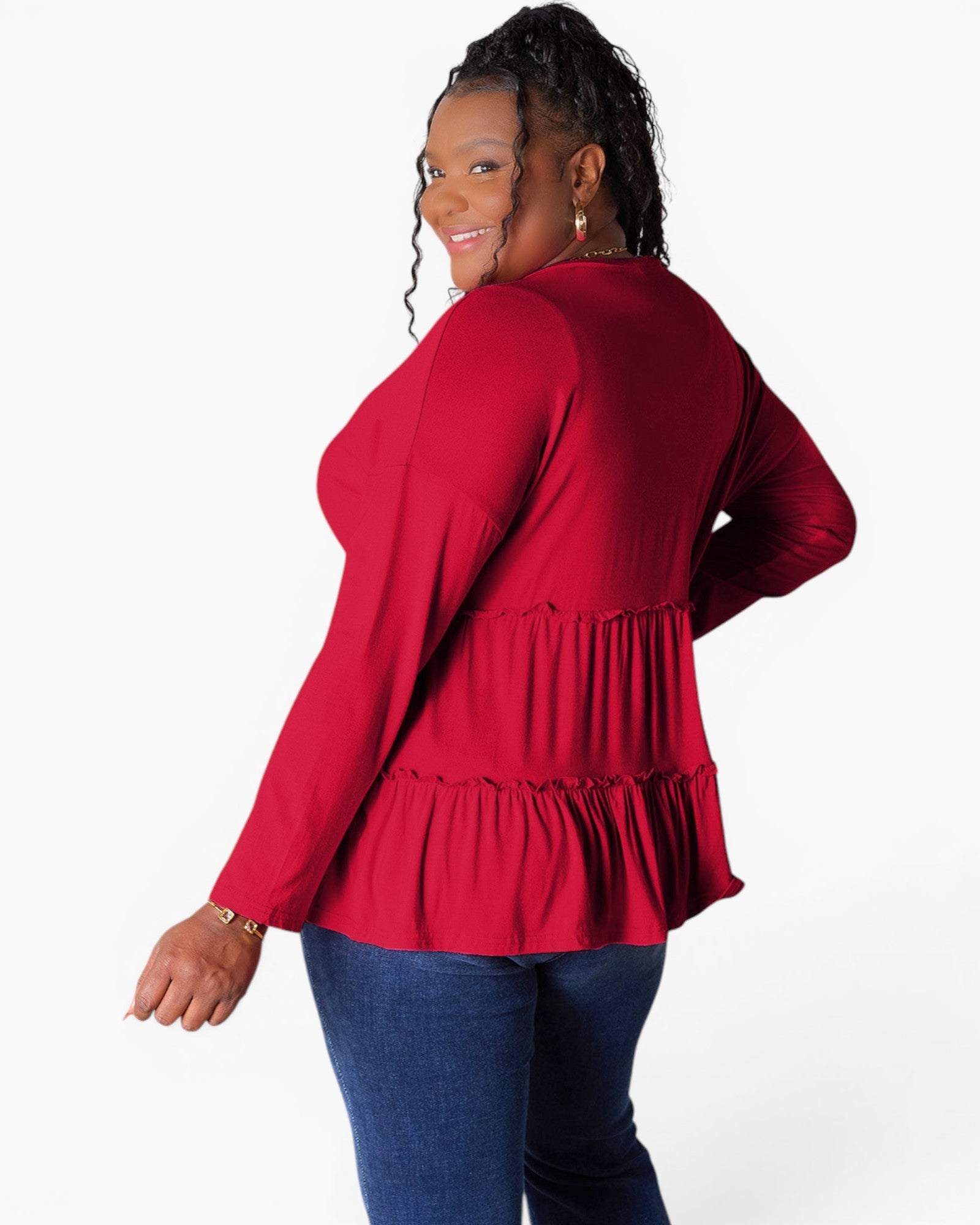 A woman modeling a red long-sleeve top with a tiered ruffle hem, shown from a back-three-quarter angle against a plain white studio background.