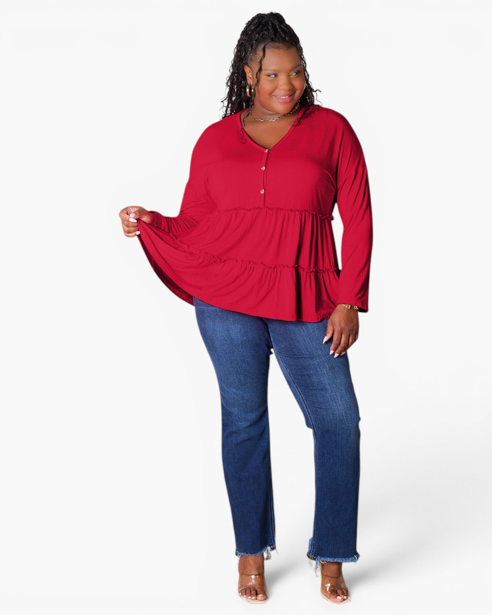 A woman modeling a red long-sleeve peplum top with a buttoned henley neckline, paired with blue jeans and clear high-heeled sandals, against a plain white studio background.