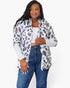 A woman modeling a white long-sleeve button-up shirt with an all-over leopard print pattern in brown and black, worn open over a white crop top and blue jeans, against a plain white background.