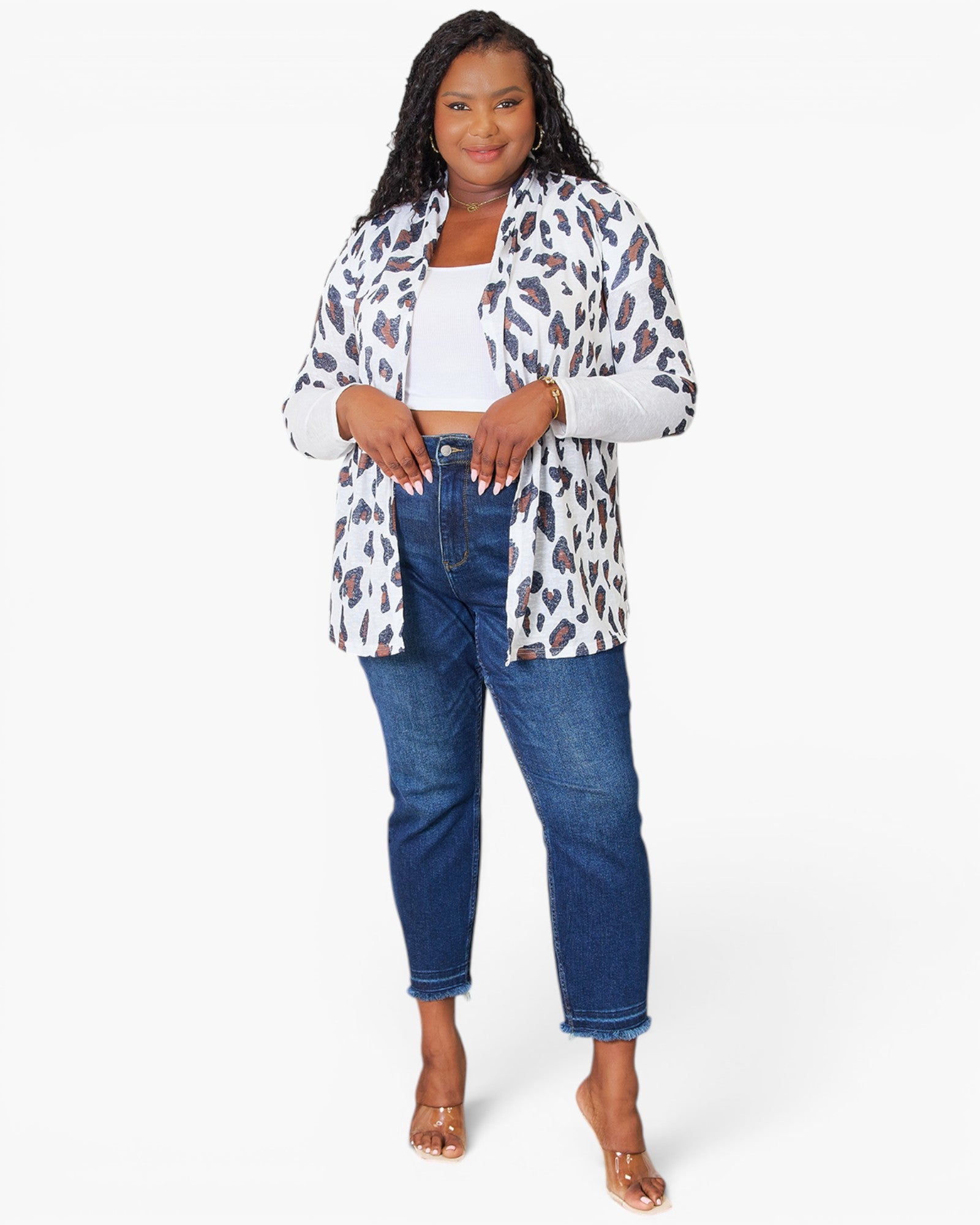 A woman modeling a white long-sleeve button-up shirt with a brown and black leopard print pattern, worn open over a white crop top and blue jeans, against a light gray studio background.