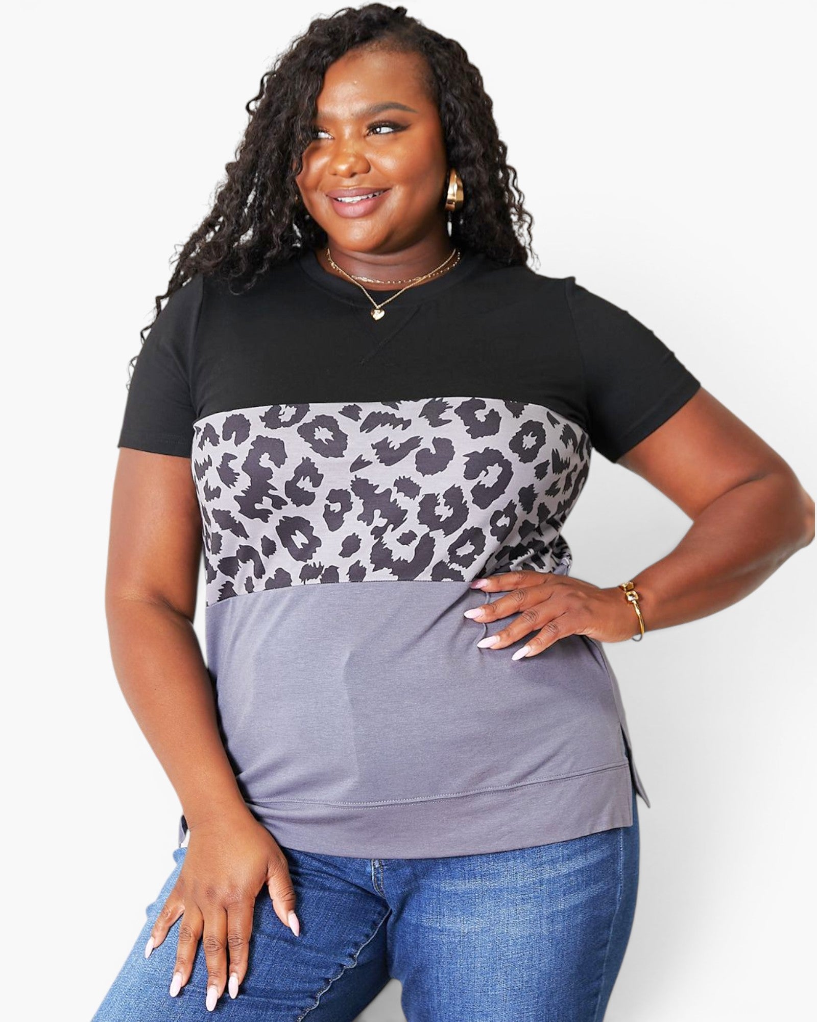 A woman modeling a short-sleeve color-block top with a black upper section, a gray and black leopard print middle band, and a muted purple-gray lower section, paired with blue jeans against a light gray studio background.