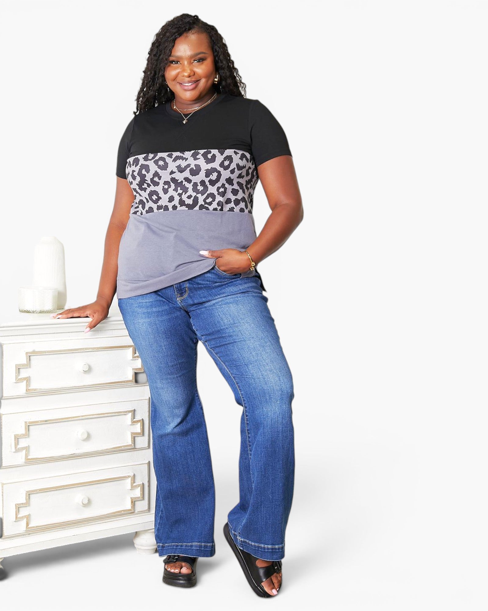 A woman modeling a short-sleeve color-block t-shirt with a black top section, a leopard print middle band, and a light gray bottom section, paired with blue jeans and black sandals, standing next to a white dresser against a white background.