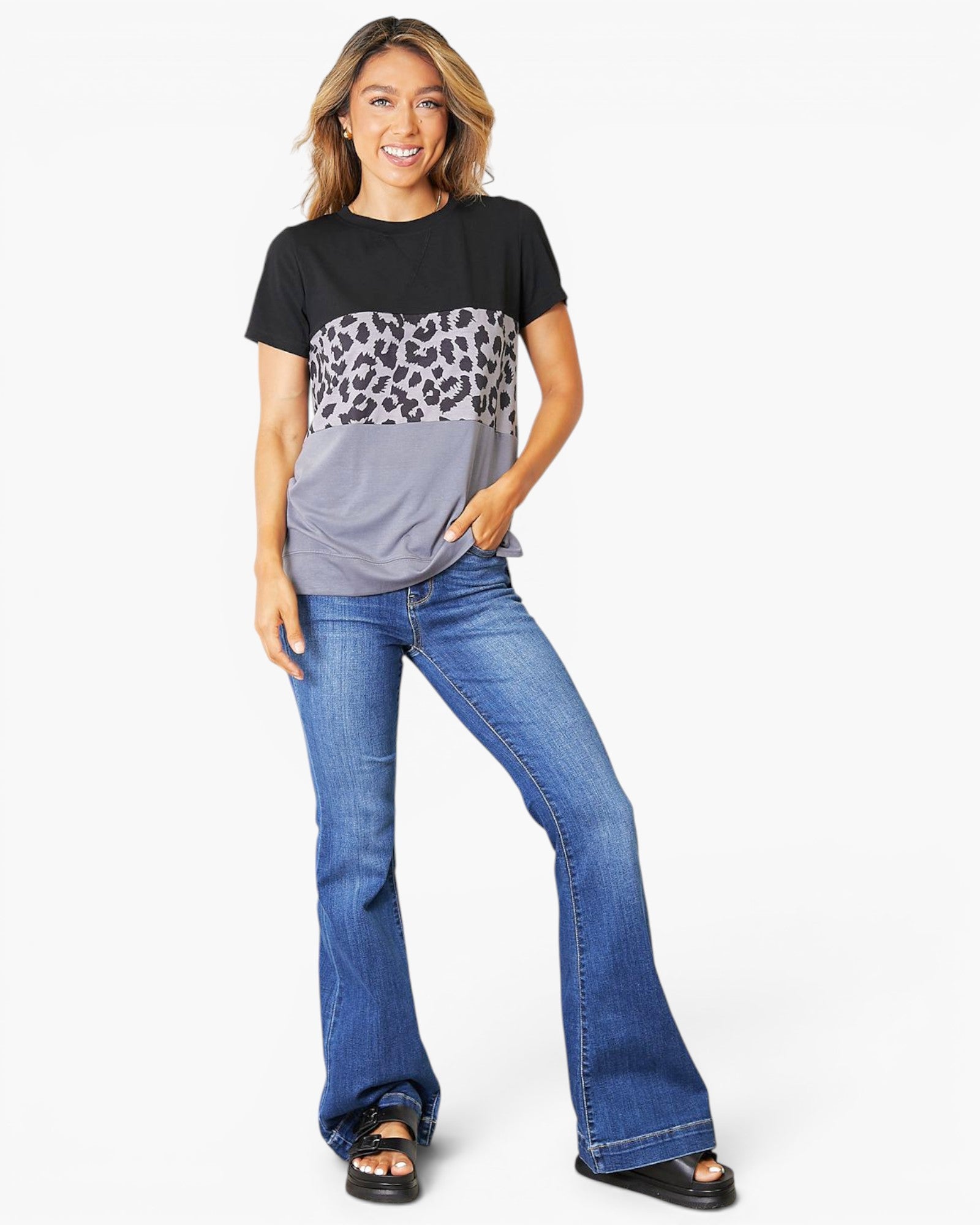 A woman modeling a short-sleeve colorblock t-shirt with a black top, leopard print middle, and gray bottom, paired with blue flare jeans and black sandals, against a plain white background.