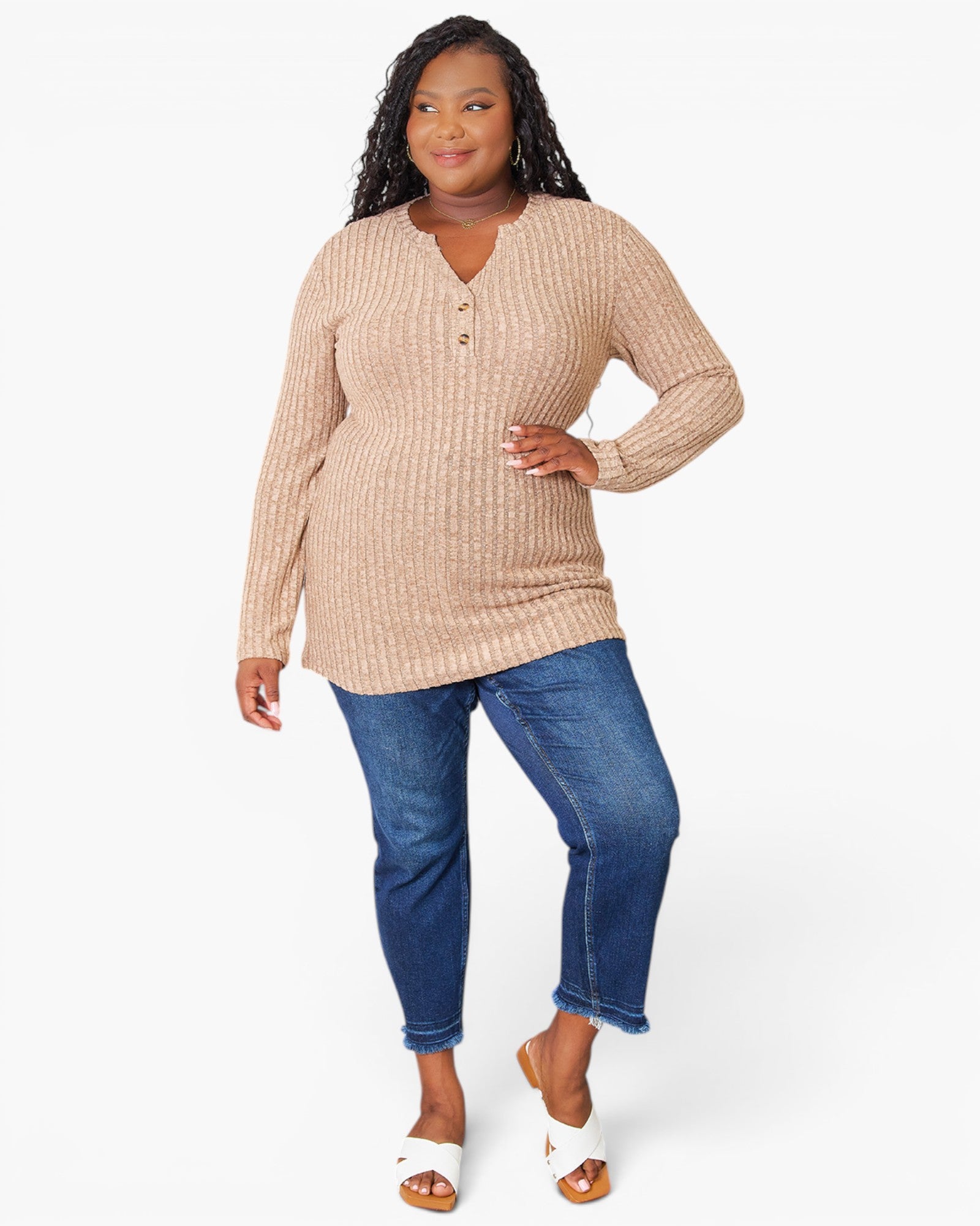 A woman modeling a beige ribbed knit long-sleeve henley top with blue jeans and white slide sandals, photographed against a clean white studio background.
