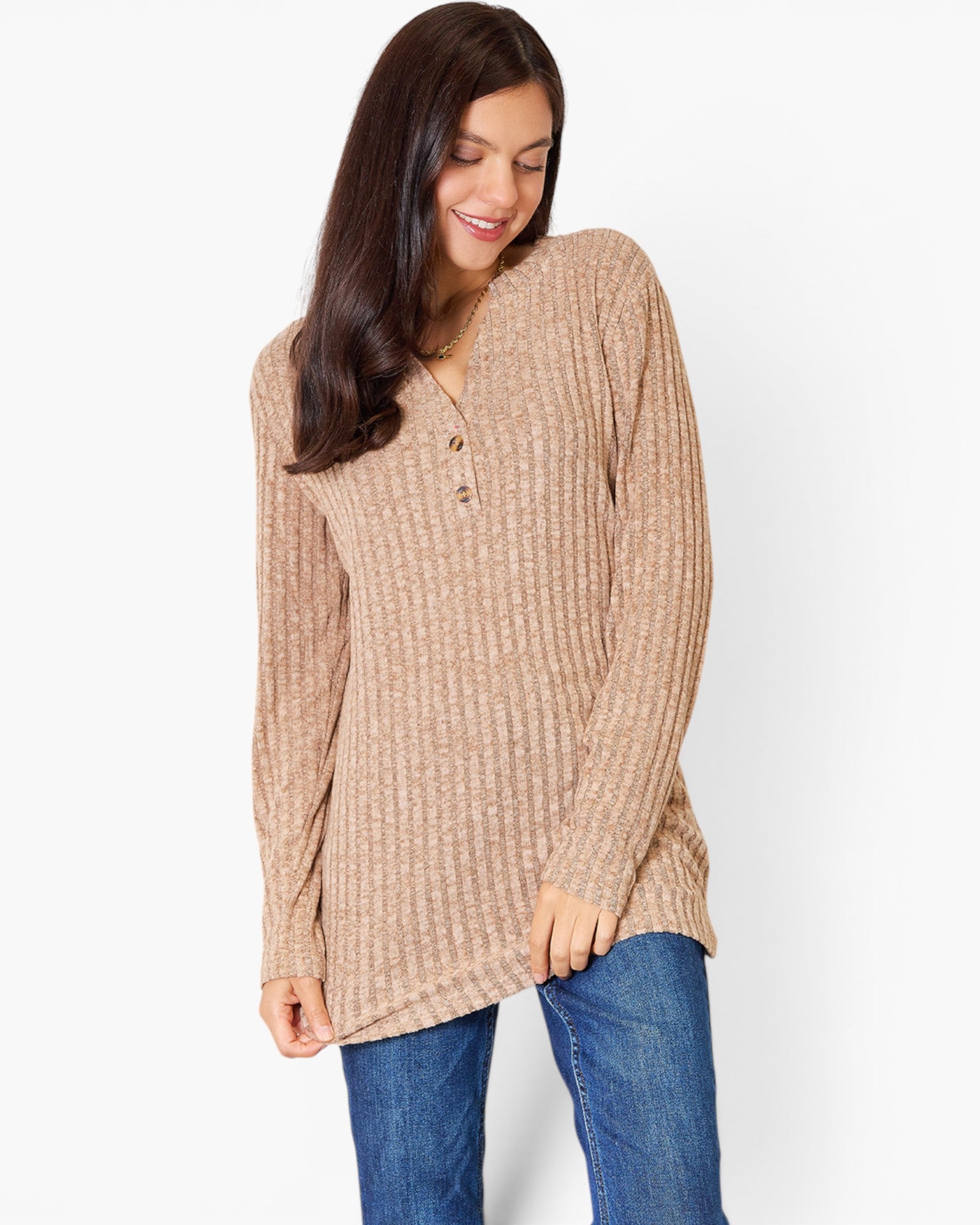 A woman wearing a beige ribbed knit long-sleeve tunic sweater with a buttoned henley neckline, paired with blue jeans, against a plain white studio background.