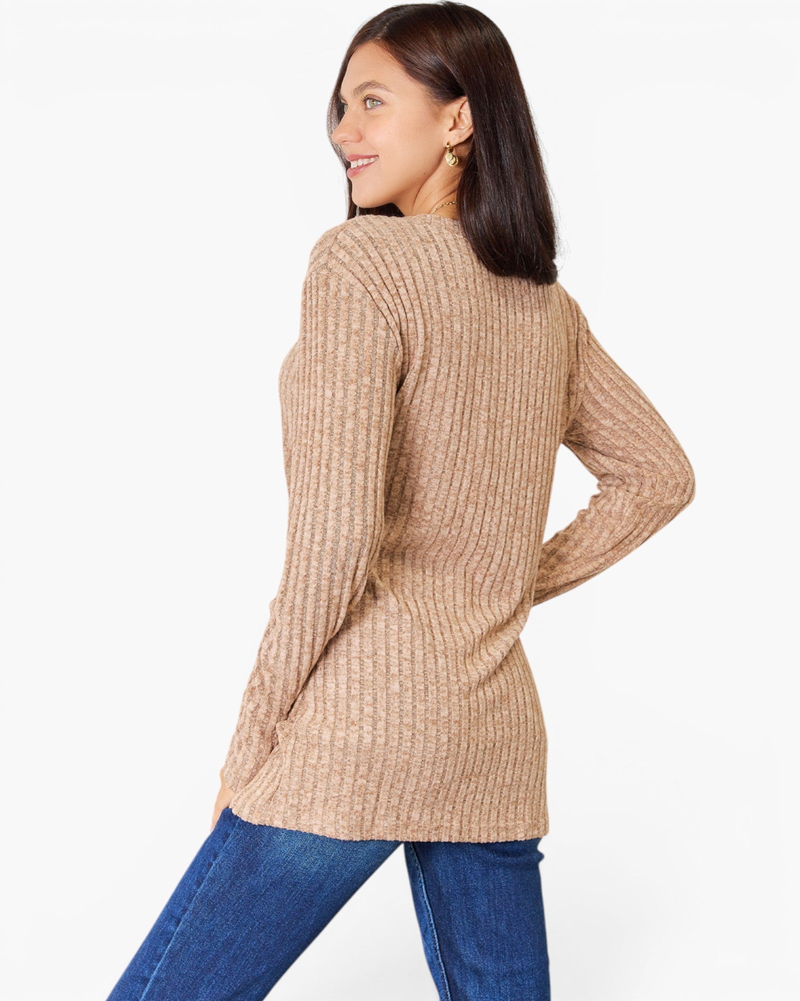 A woman wearing a beige ribbed knit long-sleeve tunic sweater and blue jeans, shown from a three-quarters back view against a plain white studio background.