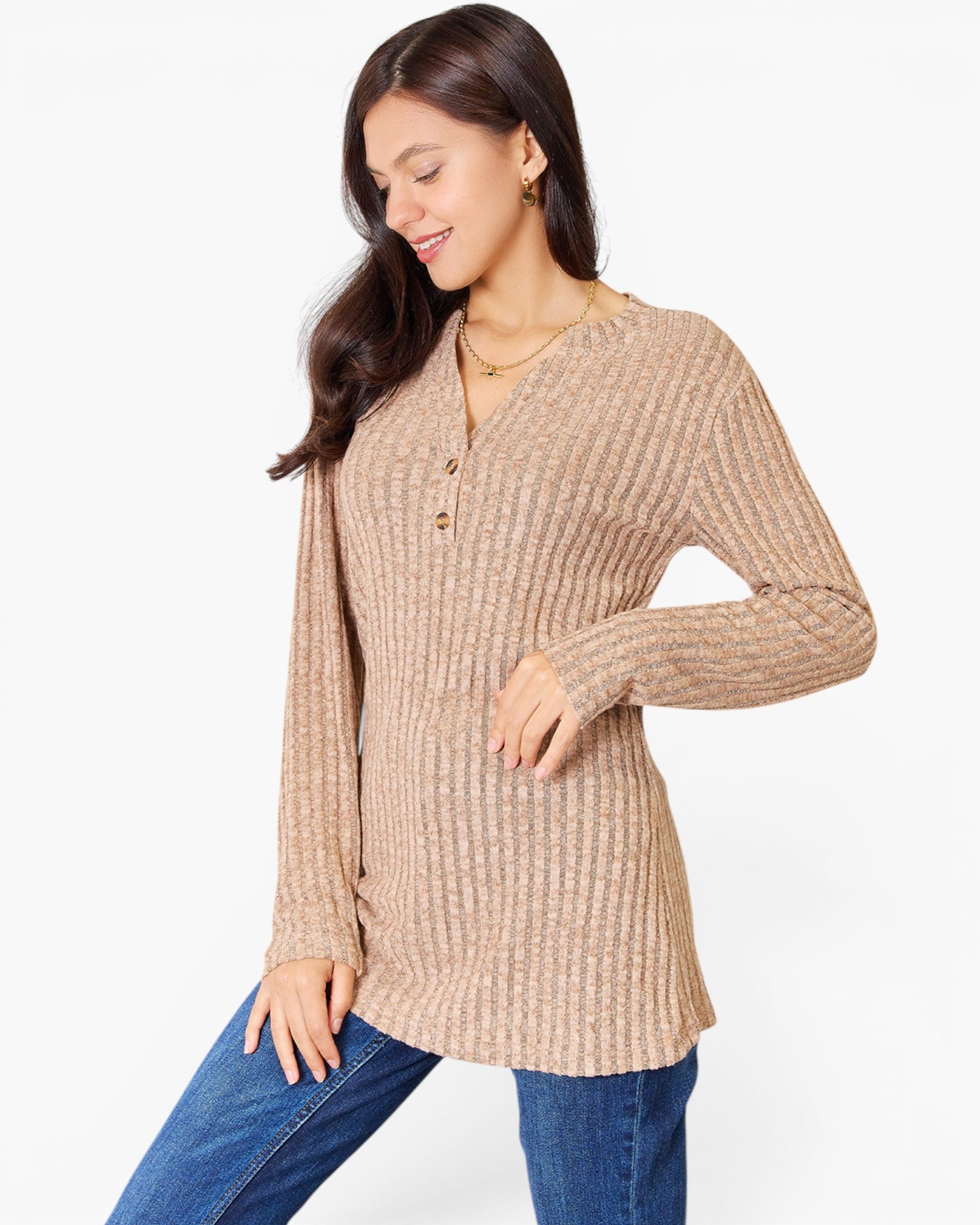 A woman wearing a beige ribbed knit long-sleeve tunic top with a buttoned henley neckline, paired with blue jeans, against a plain white studio background.