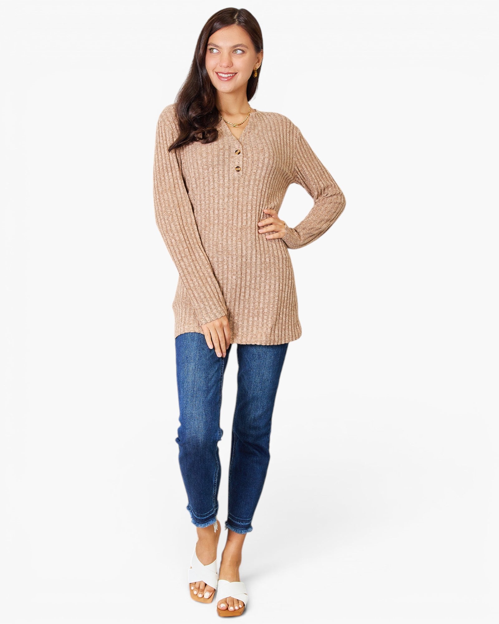 A woman modeling a beige ribbed knit long-sleeve henley tunic top with blue jeans and white slide sandals, standing against a plain white studio background.