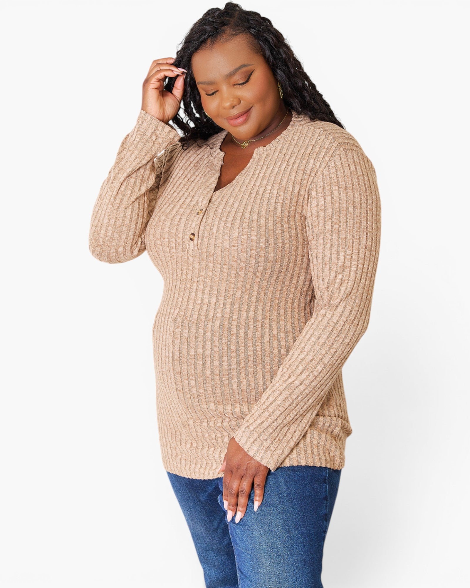 A woman wearing a beige ribbed knit long-sleeve henley top with blue jeans, standing against a plain white studio background.