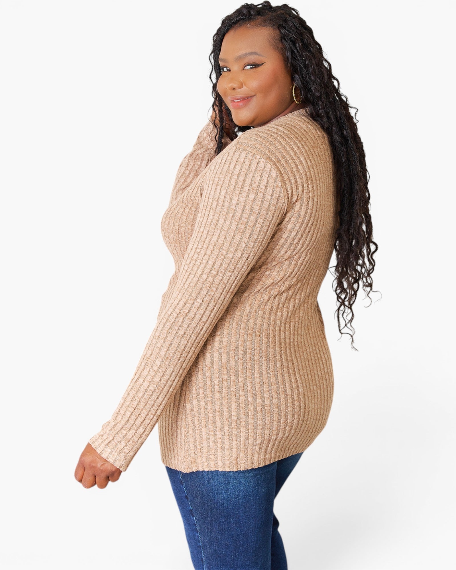 A woman modeling a beige ribbed knit sweater with blue jeans, shown from a side-back angle against a plain white studio background.