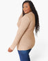 A woman modeling a beige ribbed knit sweater with blue jeans, shown from a side-back angle against a plain white studio background.