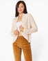 A woman modeling a light beige textured knit cardigan over a white top and brown corduroy pants, photographed against a clean white studio background.