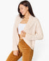A woman wearing a cream cable-knit open-front cardigan over a white tube top and brown corduroy pants, photographed against a clean white studio background.