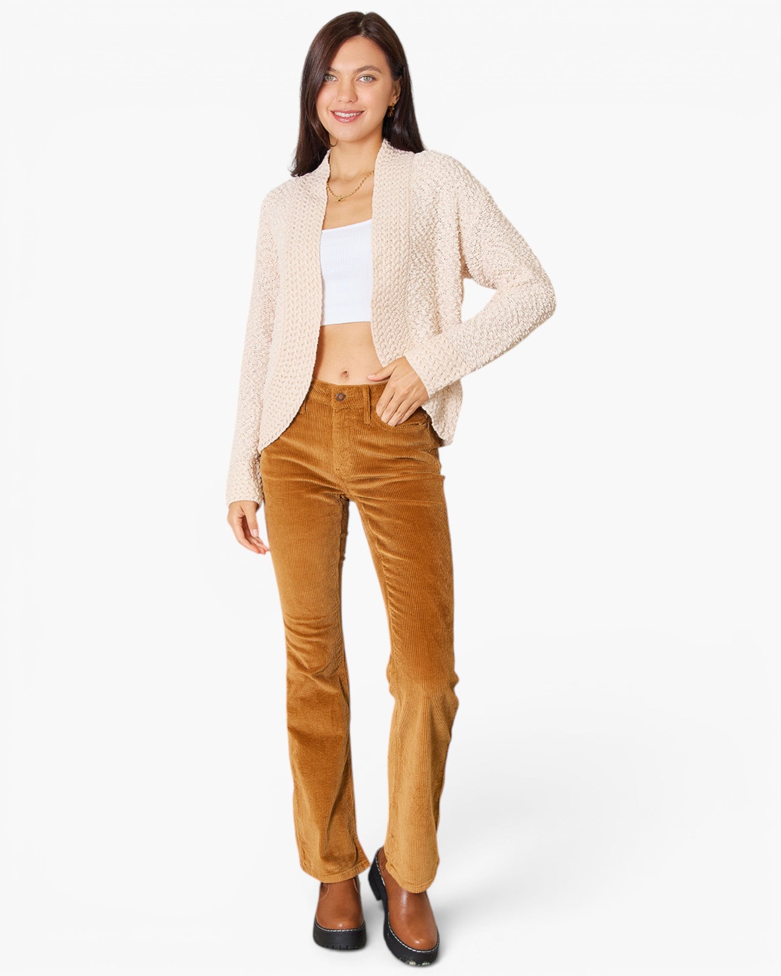 A woman modeling a cream textured knit cardigan over a white crop top and brown corduroy flare pants, paired with brown shoes, against a plain white studio background.