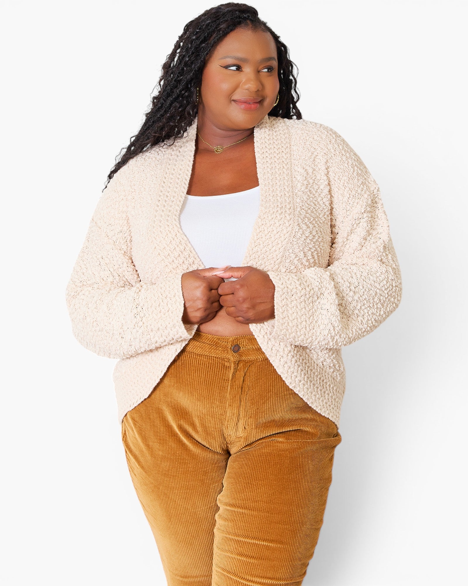 A woman modeling a cream-colored, textured boucle cardigan over a white tank top and mustard yellow corduroy pants, shot against a plain white studio background.