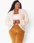 A woman modeling a cream-colored, textured boucle cardigan over a white tank top and mustard yellow corduroy pants, shot against a plain white studio background.
