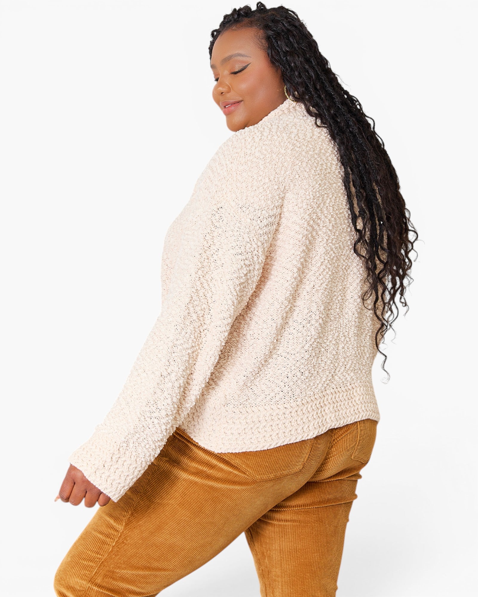 A woman wearing a cream-colored textured knit sweater and mustard brown corduroy pants, shown from a side-back angle against a plain white background.