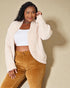 Woman wearing a cream sherpa jacket over a white top and brown pants on a beige background