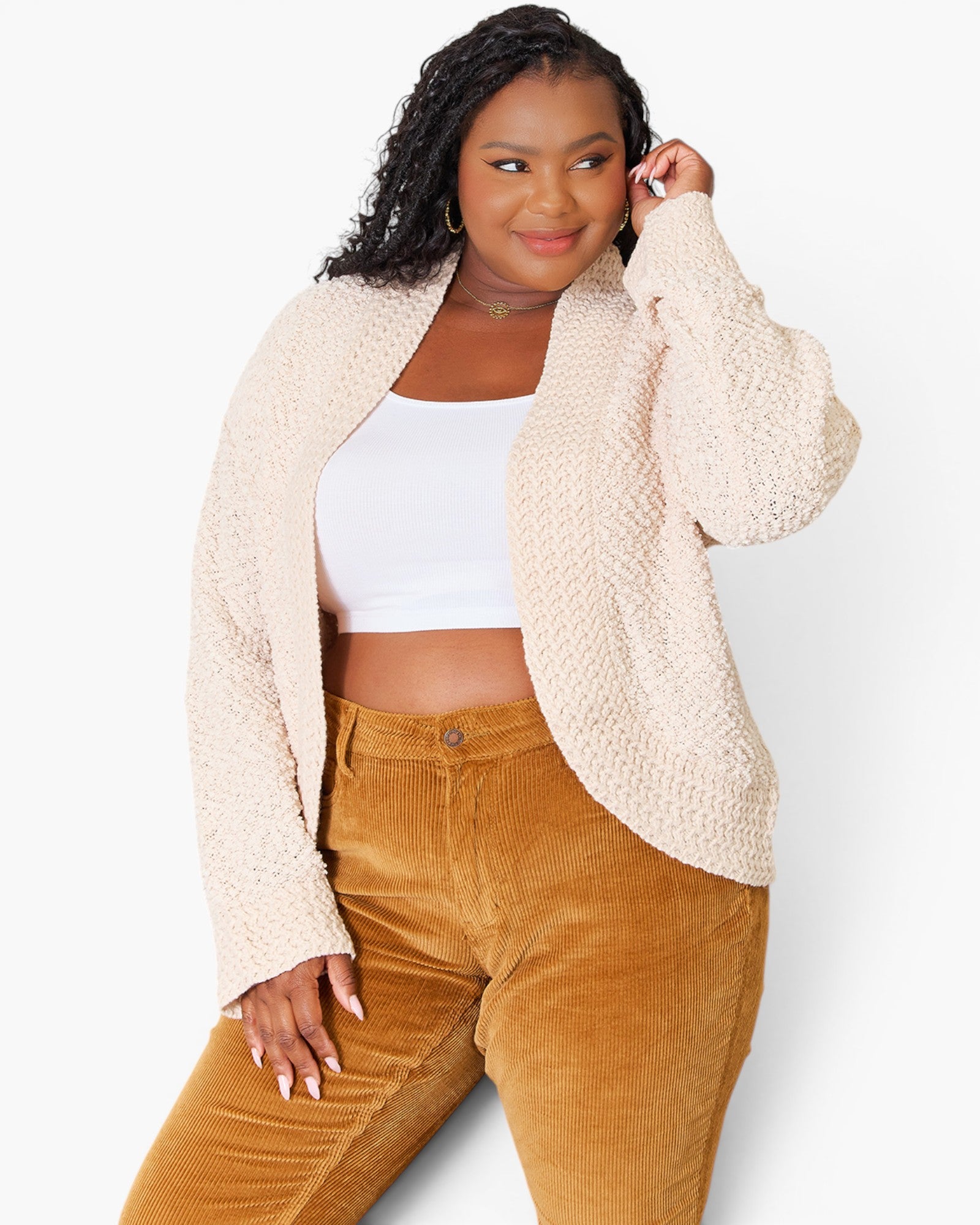 A woman modeling a cream-colored, textured boucle cardigan over a white crop top and brown corduroy pants, photographed against a clean white studio background.