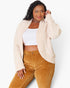 A woman modeling a cream-colored, textured boucle cardigan over a white crop top and brown corduroy pants, photographed against a clean white studio background.