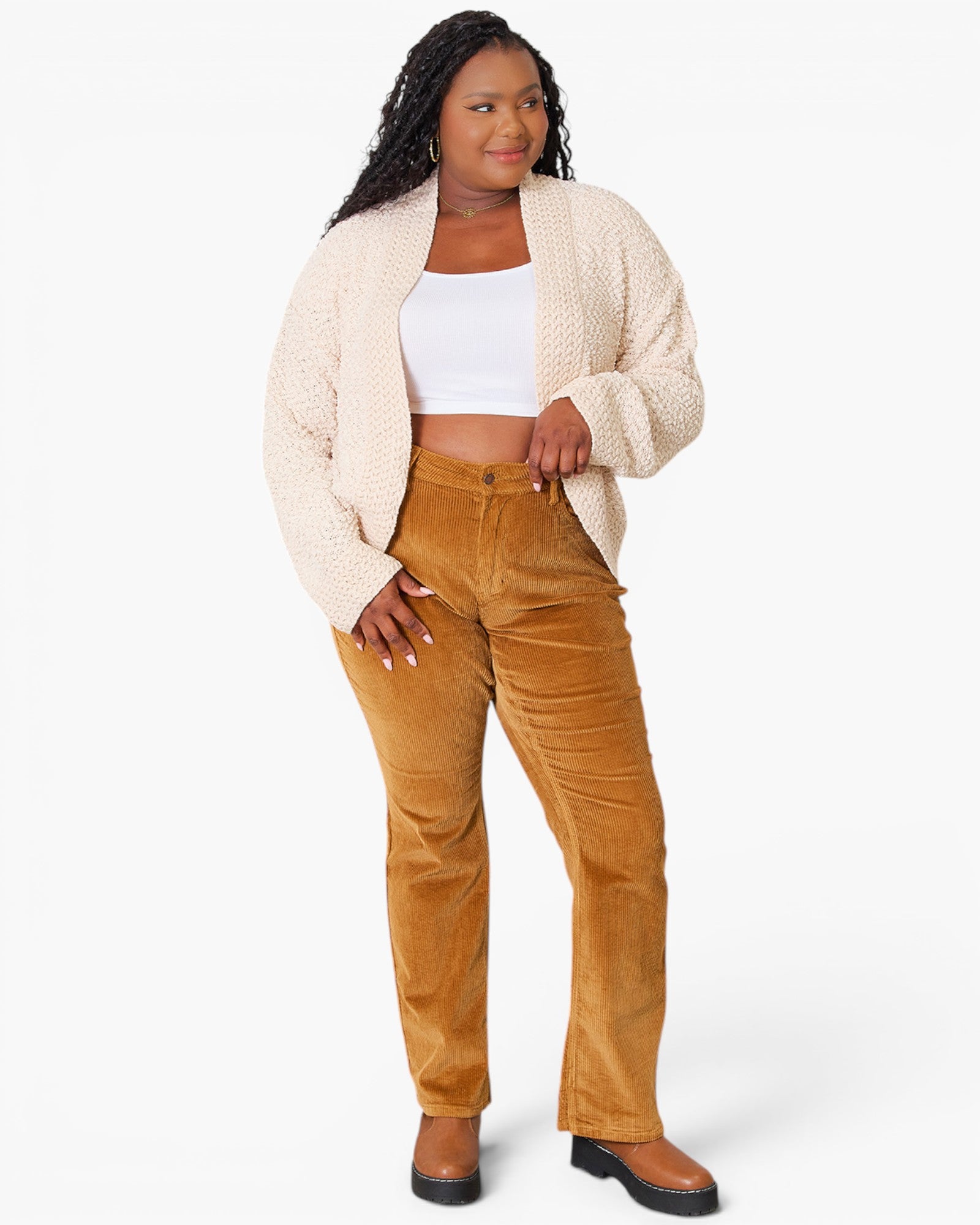 A woman modeling a cream cable-knit cardigan over a white crop top and high-waisted mustard-brown corduroy pants, paired with brown boots, against a light gray studio background.