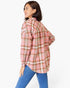 A woman wearing a pink, brown, and beige plaid flannel shirt with blue jeans, shown from a side-back angle against a plain white studio background.