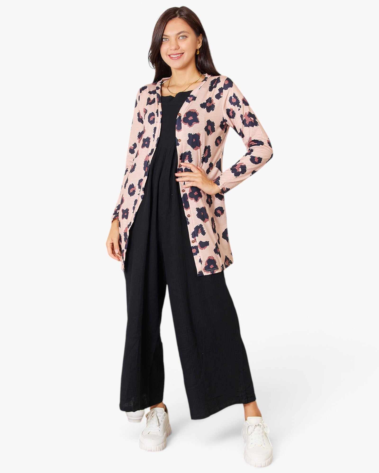 A woman modeling a light pink leopard-print cardigan over a black jumpsuit, paired with white sneakers, against a plain white studio background.