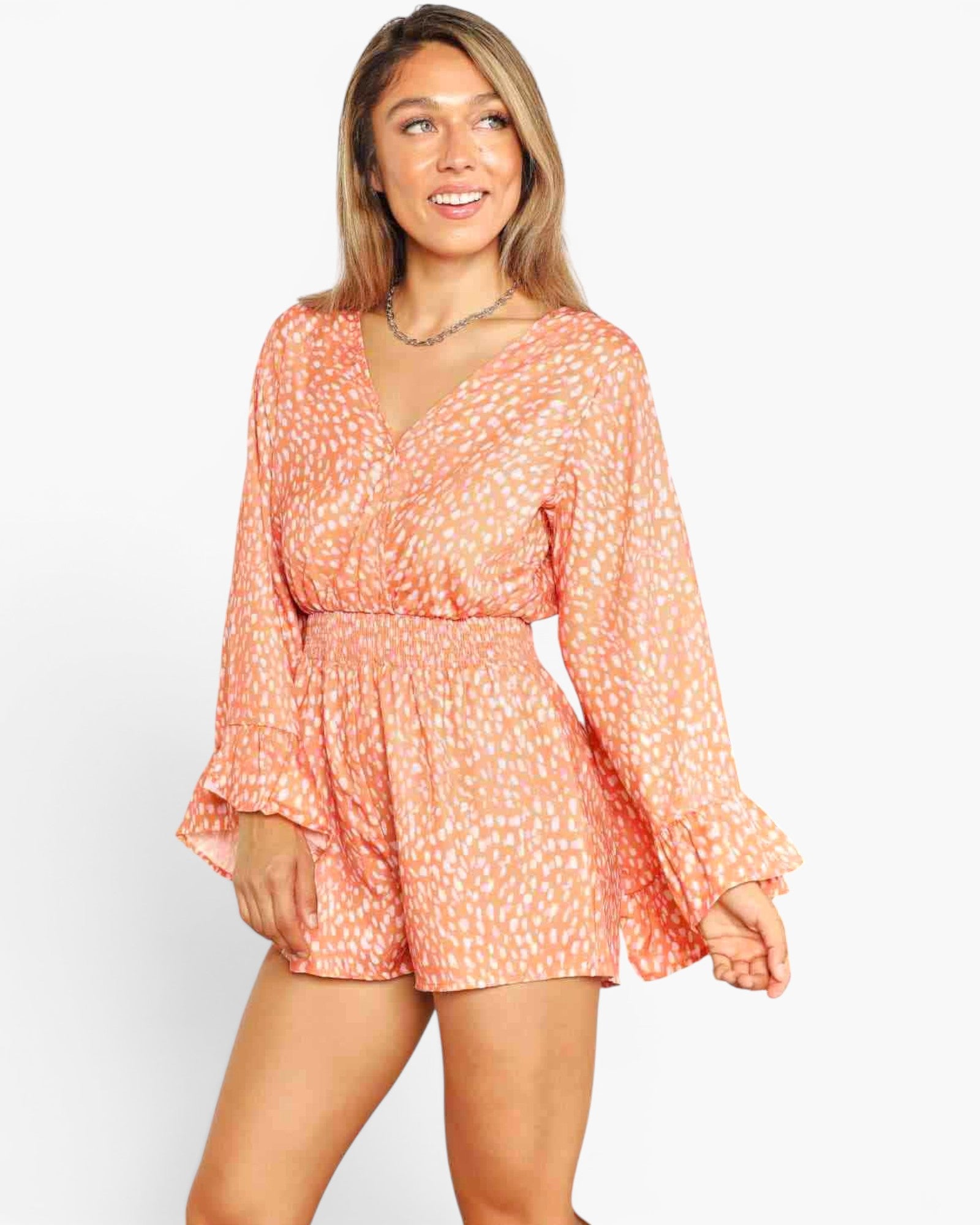 A woman modeling a peach and white patterned long-sleeve romper with a V-neckline and flared cuffs against a plain white background.