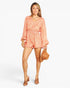 A woman modeling a peach-orange and white patterned romper with long bell sleeves and a V-neckline, holding a brown hat against a light gray studio background.