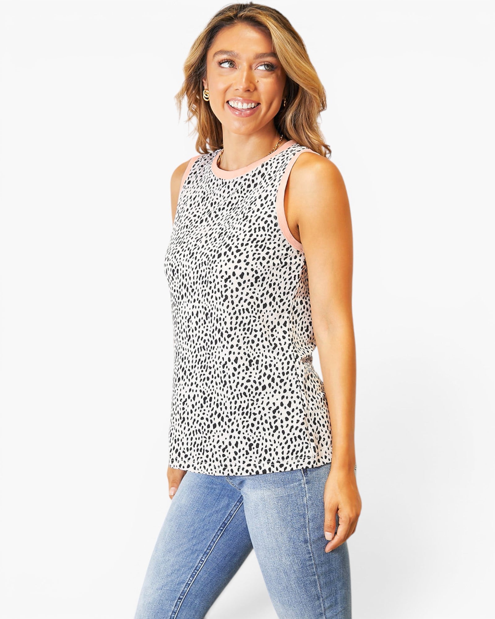 A woman modeling a sleeveless white tank top with a black dalmatian-style spot pattern, paired with light blue jeans, against a plain white studio background.