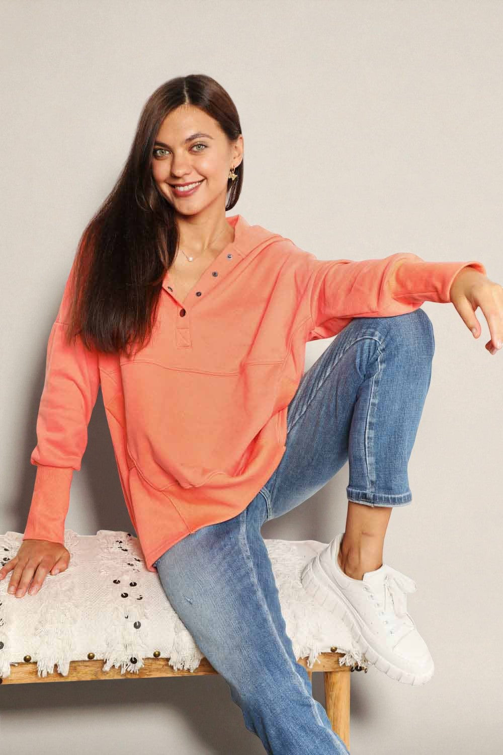 Woman wearing a coral sweatshirt and blue jeans sitting on a white surface.