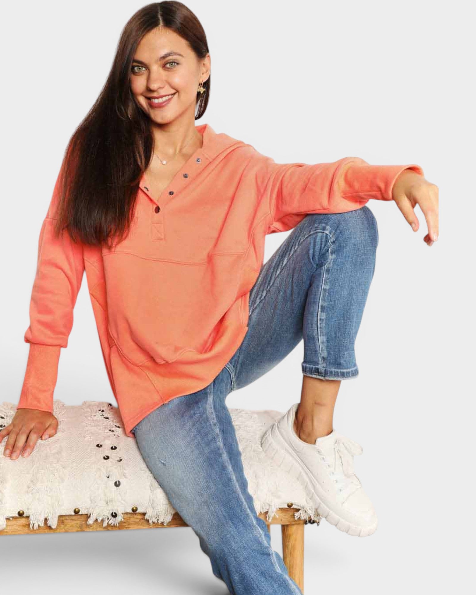 Woman wearing a coral long-sleeve top and blue jeans sitting on a white stool.