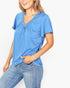 A woman wearing a heather blue short-sleeve V-neck top with a gathered front detail and button accent, paired with light-wash blue jeans, against a plain light background.