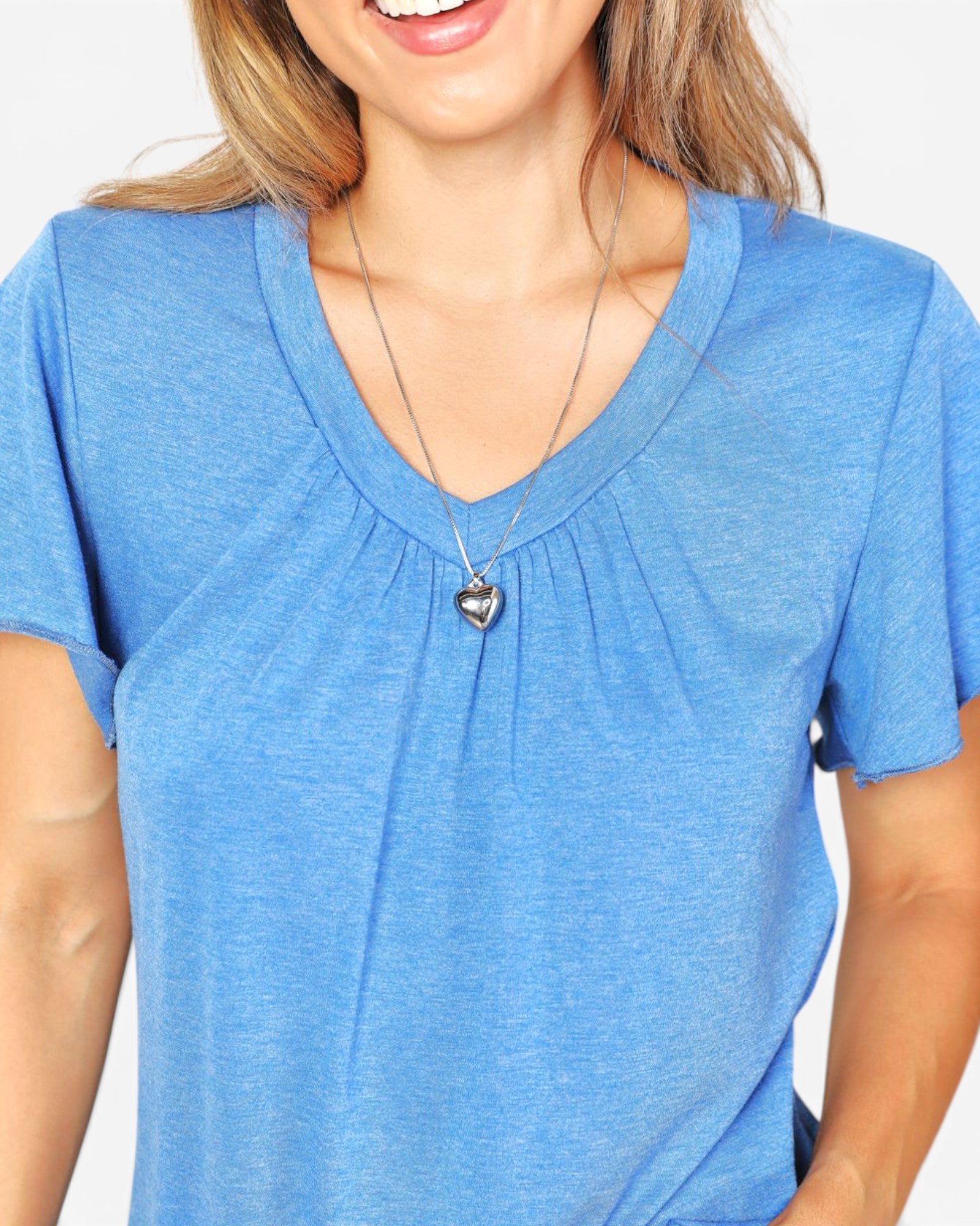A woman wearing a heathered blue short-sleeve V-neck top with a gathered neckline and a silver heart pendant necklace, shown against a plain light background.