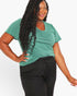 A woman modeling a short-sleeve green V-neck t-shirt with a twisted hem detail, paired with black sweatpants, against a plain white background.