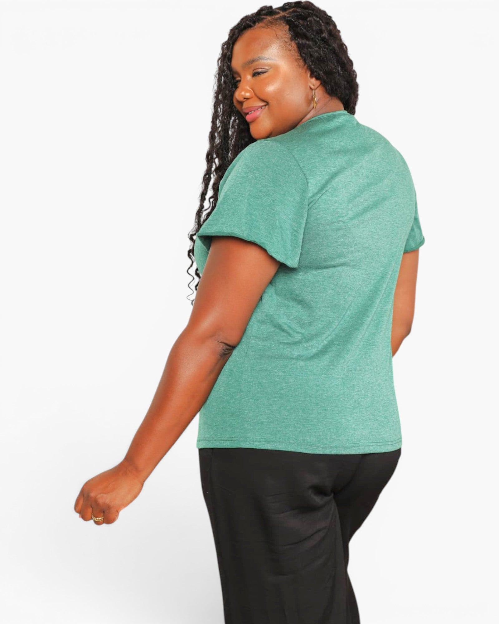 A woman wearing a heathered sage green short-sleeve t-shirt and black pants, shown from a side-back angle against a plain white studio background.